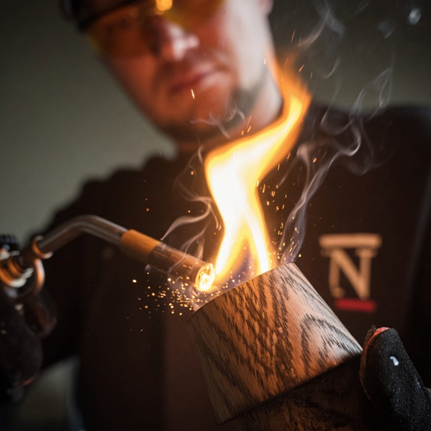 A craftsperson using a torch to char the inside of an oak whiskey tumbler, with the flame visible above the rim