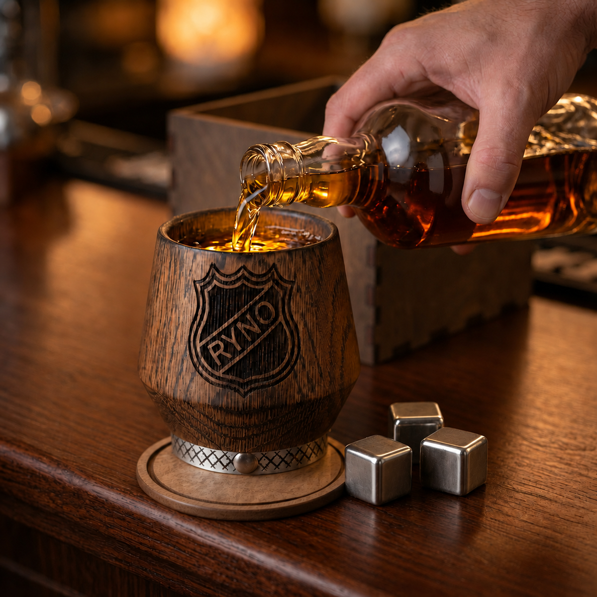 A hand pours amber whiskey into a wooden tumbler engraved with a RYNO shield. The tumbler rests on a round wooden coaster beside three stainless steel chilling cubes, with a wooden gift box behind.