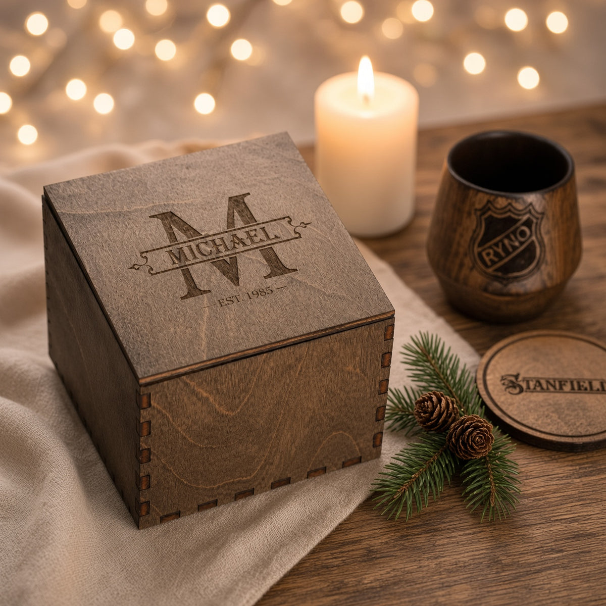 Closed wooden gift box with an engraved decorative M monogram on the lid. A wooden whiskey tumbler and part of a personalized round coaster sit beside it on a neutral surface.