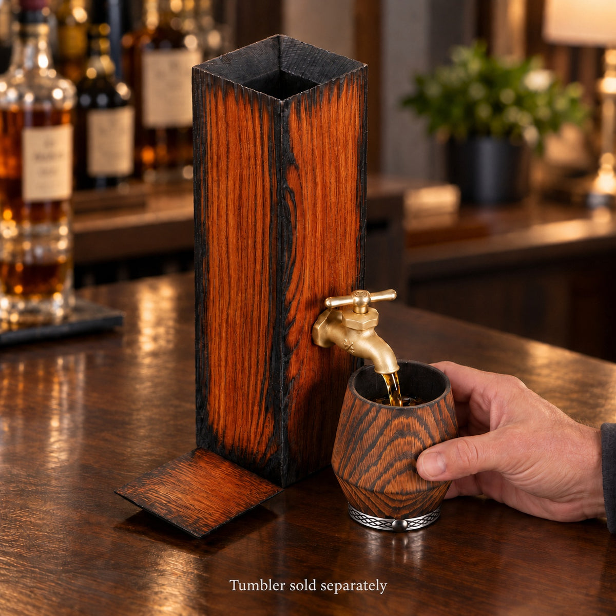 Ember Oak whiskey dispenser on a home bar with a hand holding a wooden tumbler beneath the brass tap. Tumbler sold separately.