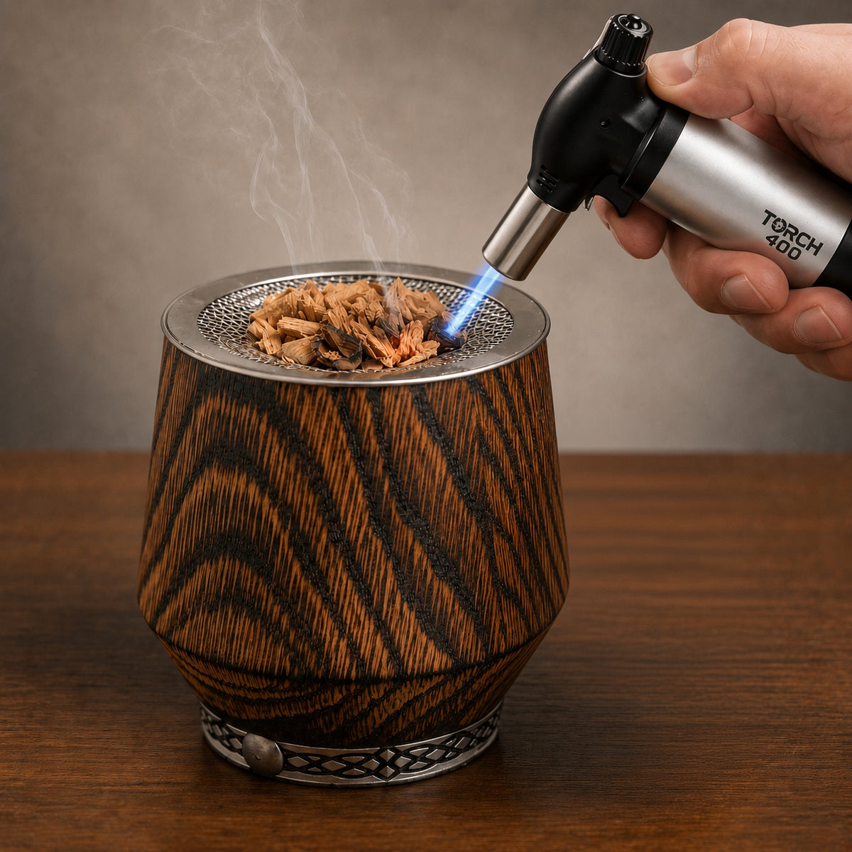 A handheld torch lighting wood shavings in a metal mesh resting at the rim of a charred oak whiskey tumbler.